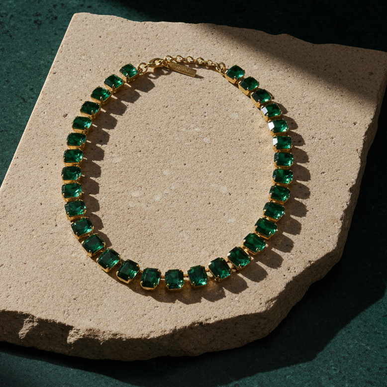 Single-strand necklace of emerald-cut green crystals in gold-tone settings, laid on a stone slab with its extension chain and branded tag.