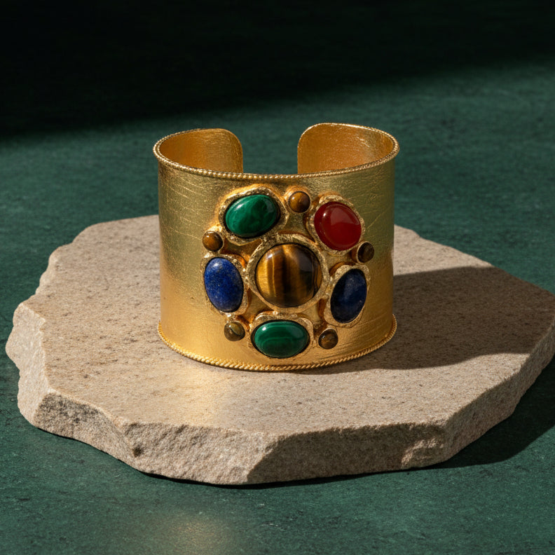 Wide brushed gold cuff standing on a pale stone slab, set with a round tiger's eye cabochon ringed by green malachite, blue lapis and red carnelian ovals in gold bezels.