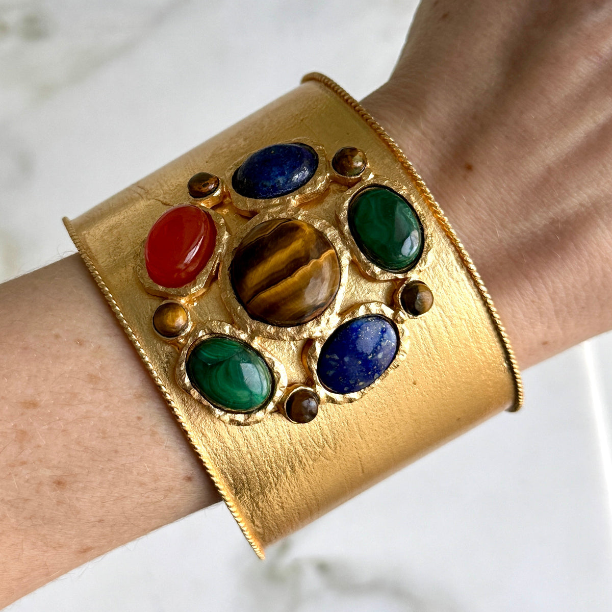 The brushed gold cuff worn on a wrist, showing the raised cluster of tiger's eye, malachite, lapis and carnelian cabochons across the front.
