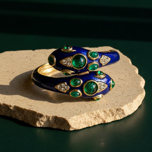 Cobalt blue enamel double-headed snake cuff on a pale stone slab, both heads set with domed green cabochons and pavé-set clear stones.