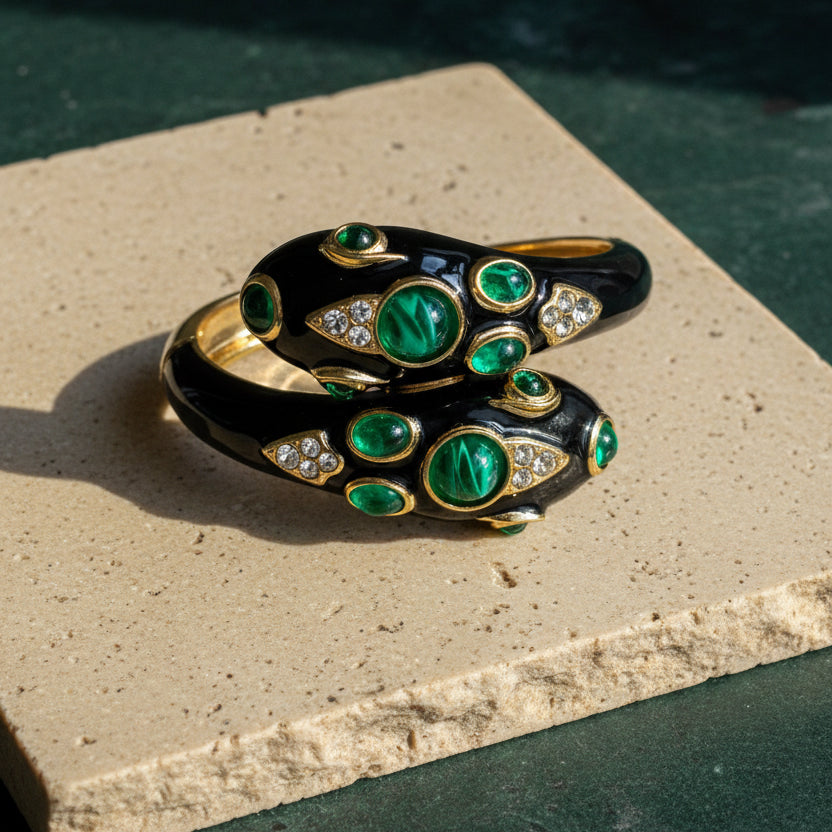 Black enamel double-headed snake cuff on a pale stone block, both heads set with domed green cabochons and pavé-set clear stones in gold bezels.