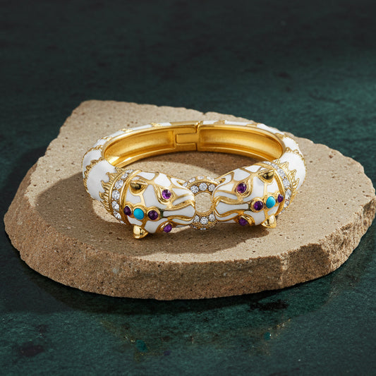 White enamel double panther cuff on a pale stone slab, two panther heads facing each other across a pavé-set link, with purple and turquoise-blue cabochons.