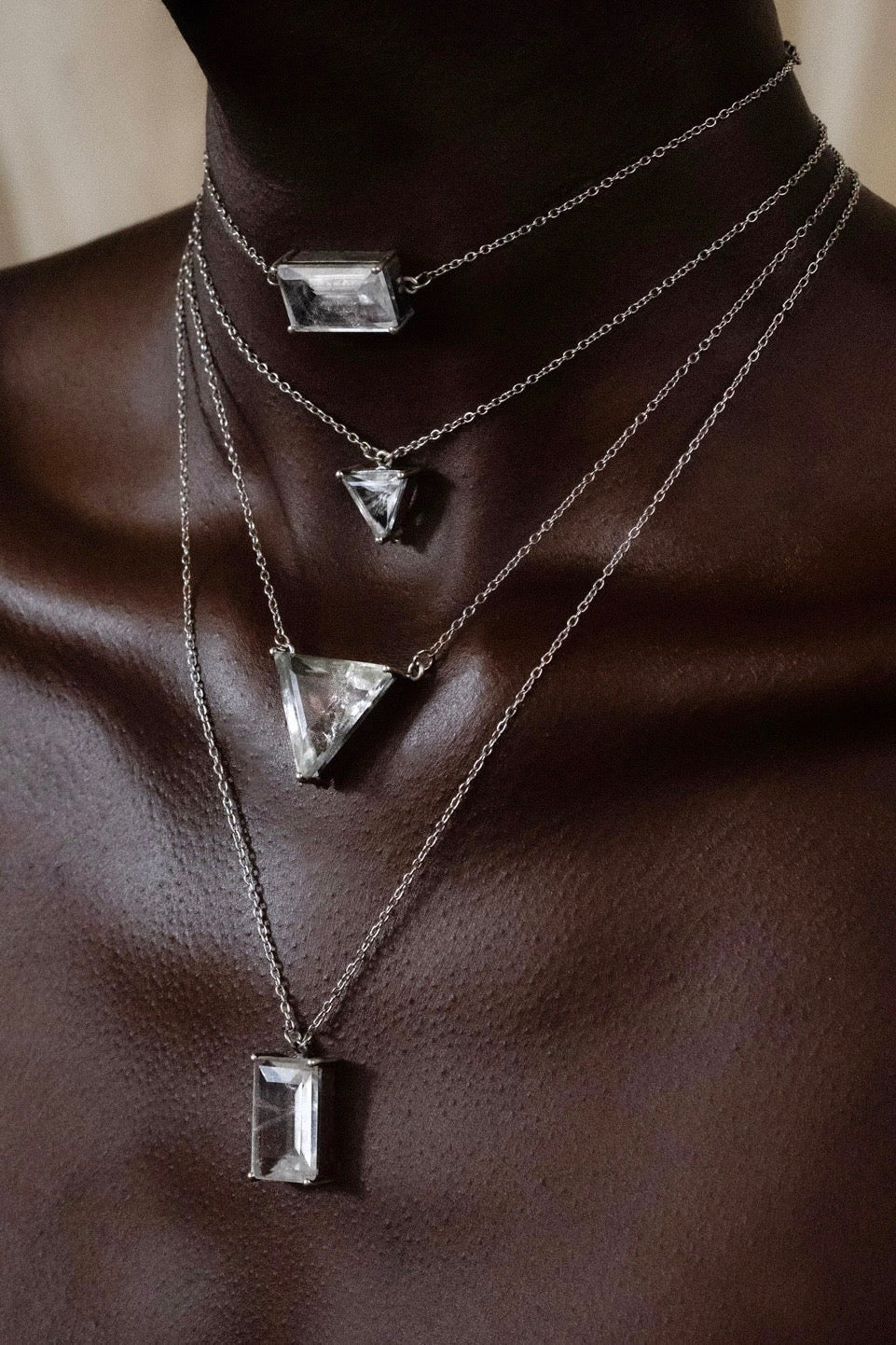 Four Obscura stacking shards layered on bare skin — emerald-cut, triangle and rectangular natural quartz pendants on fine silver chains at four descending lengths