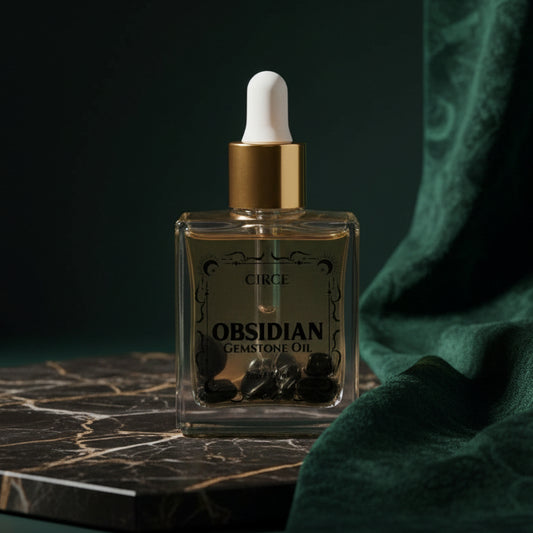 The smaller Black Obsidian gemstone oil bottle on dark marble beside green velvet, its label reading 'Obsidian Gemstone Oil', with tumbled black obsidian chips resting in the oil.