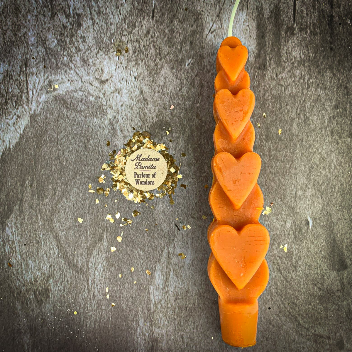 The Orange (Breakthrough) Love Spell Heart Taper in bright orange beeswax on a mottled stone backdrop, the gold-flecked Madame Pamita token beside it.