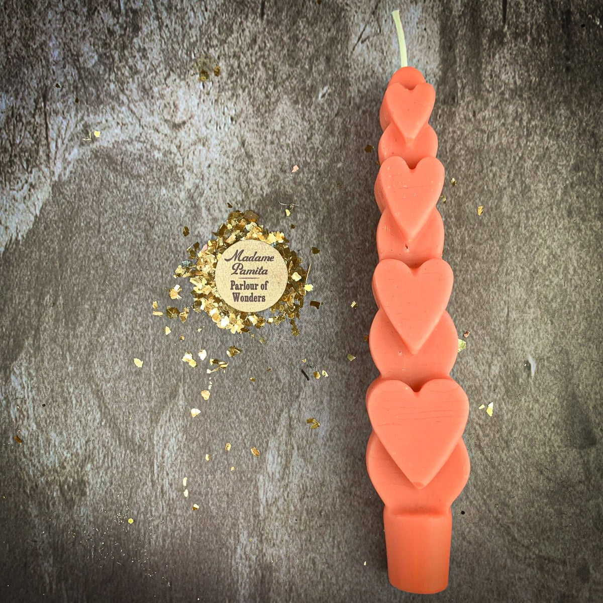 The Pink (Romance) Love Spell Heart Taper in warm coral beeswax on a mottled stone backdrop, the gold-flecked Madame Pamita token beside it.