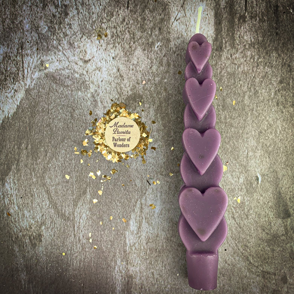 The Purple (Power) Love Spell Heart Taper in dusty mauve beeswax on a mottled stone backdrop, the gold-flecked Madame Pamita token beside it.