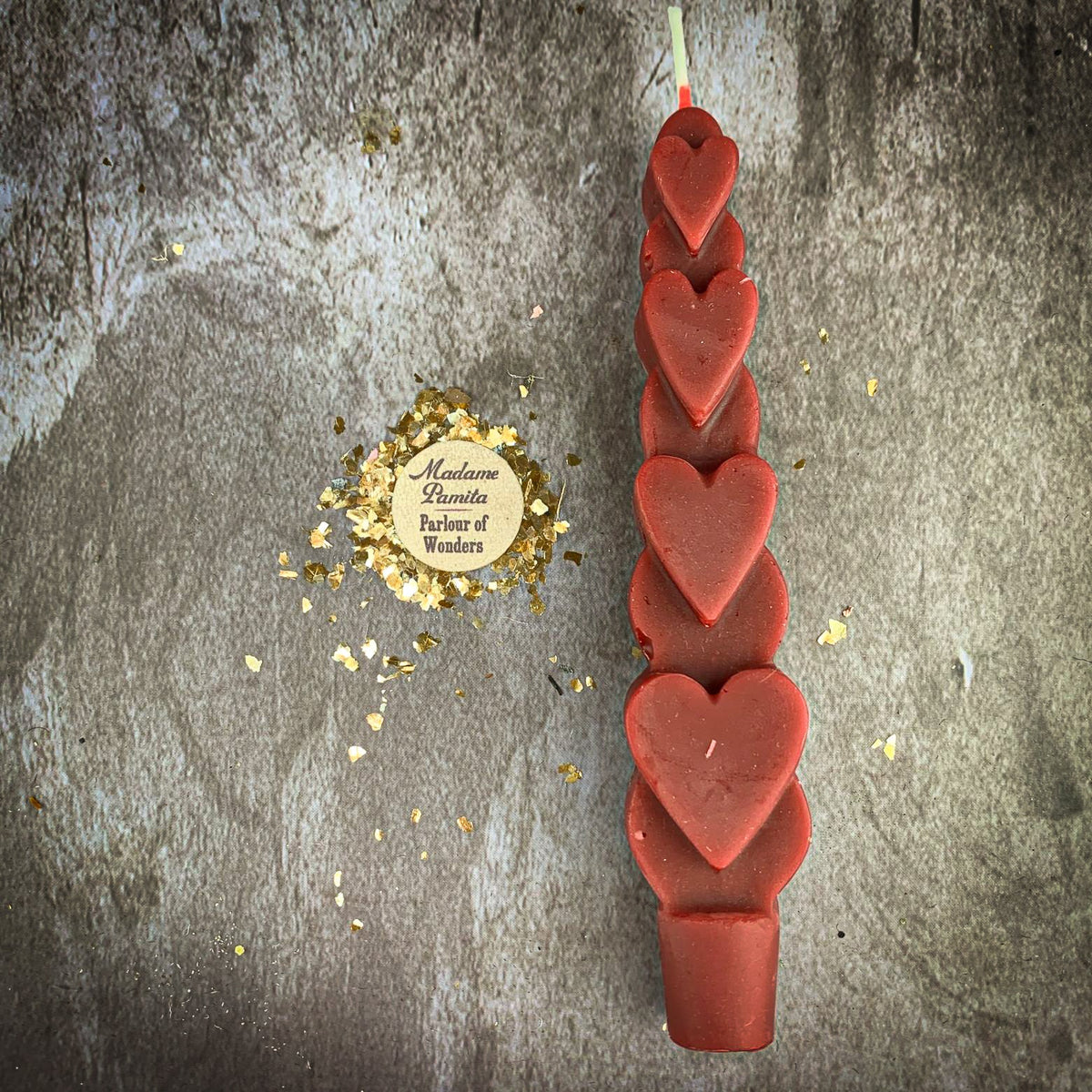 The Red (Passion) Love Spell Heart Taper in brick-red beeswax on a mottled stone backdrop, the gold-flecked Madame Pamita token beside it.