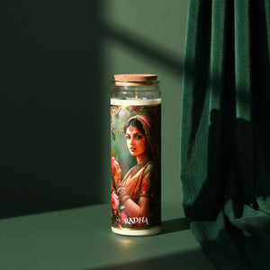 The Radha altar candle standing on a green surface beside a dark green drape, sunlight striping the wall behind; a cork lid closes the tall glass jar.