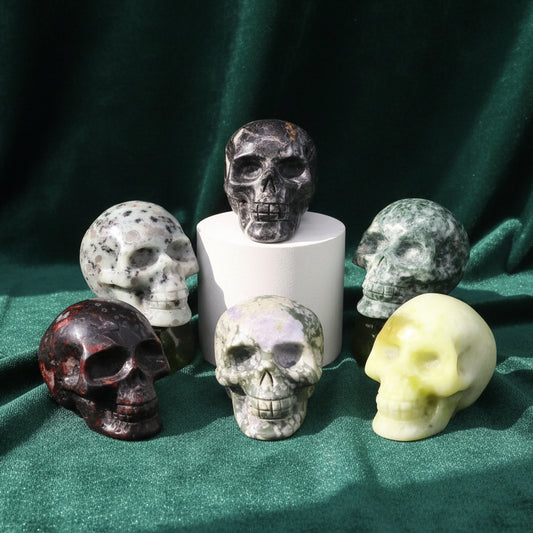 Six carved crystal skulls grouped on green velvet around a white riser, in speckled gray, mottled cream, green, dark red and pale yellow stone