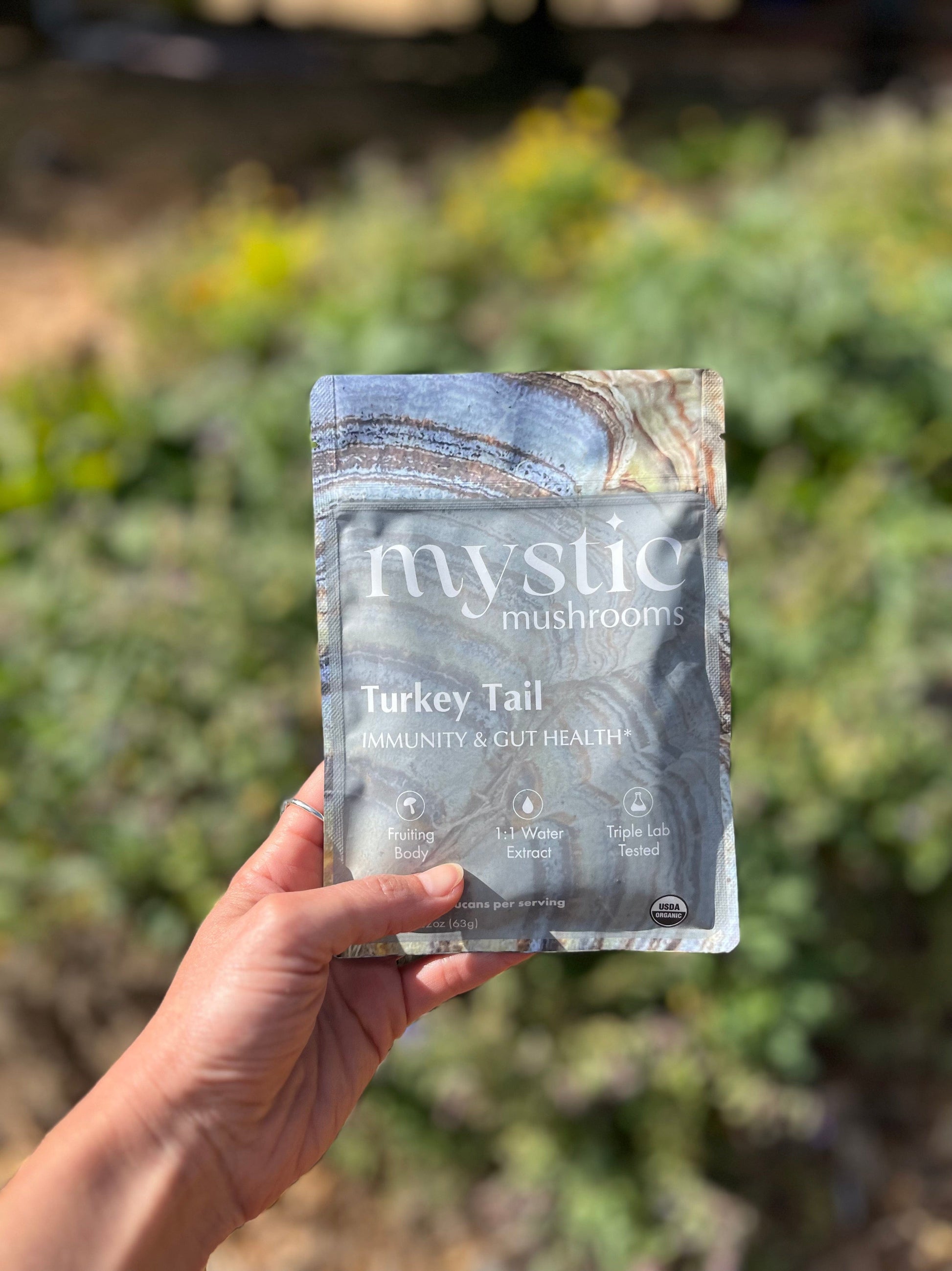 A hand holding the Mystic Mushrooms Turkey Tail pouch outdoors in front of green shrubs, showing the banded turkey tail mushroom artwork