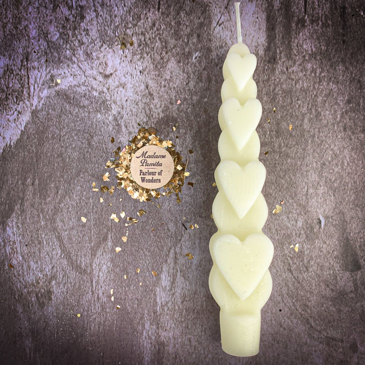 The White (Blessing) Love Spell Heart Taper in cream beeswax on a mottled stone backdrop, the gold-flecked Madame Pamita token beside it.