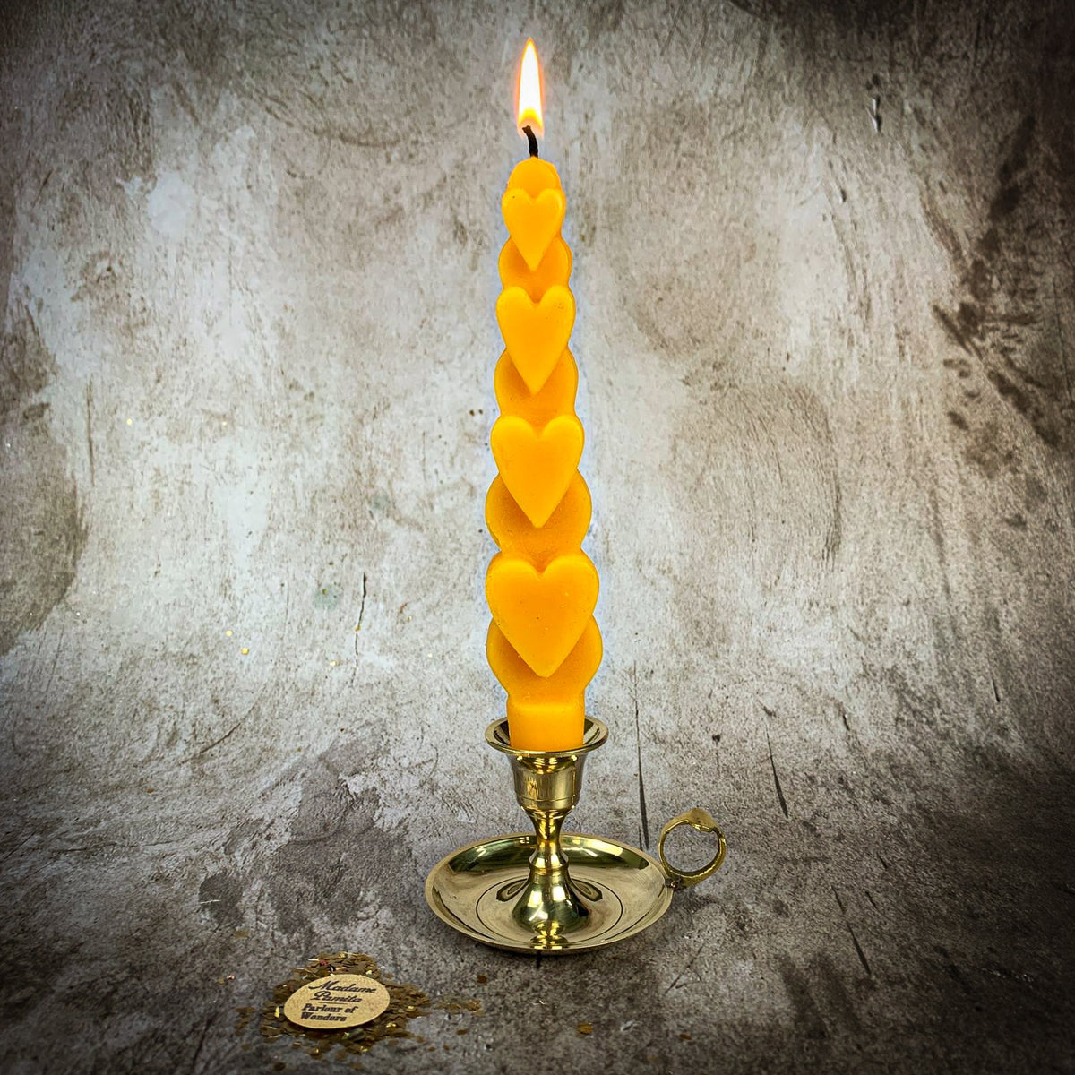 The Yellow (Happiness) Love Spell Heart Taper burning in a small brass chamberstick on a mottled stone backdrop, the Madame Pamita token resting on the floor beside it.