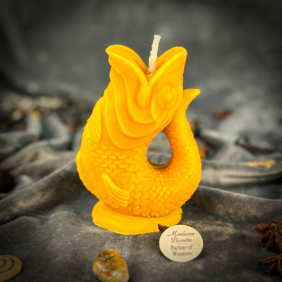 Golden yellow fish-shaped candle standing on its round foot on gray velvet, the Madame Pamita token beside it