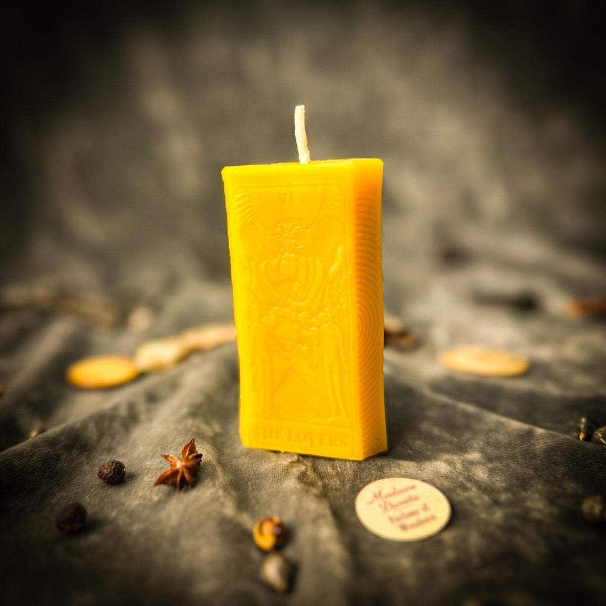 Golden yellow beeswax The Lovers tarot card candle on gray cloth with scattered coins and herbs