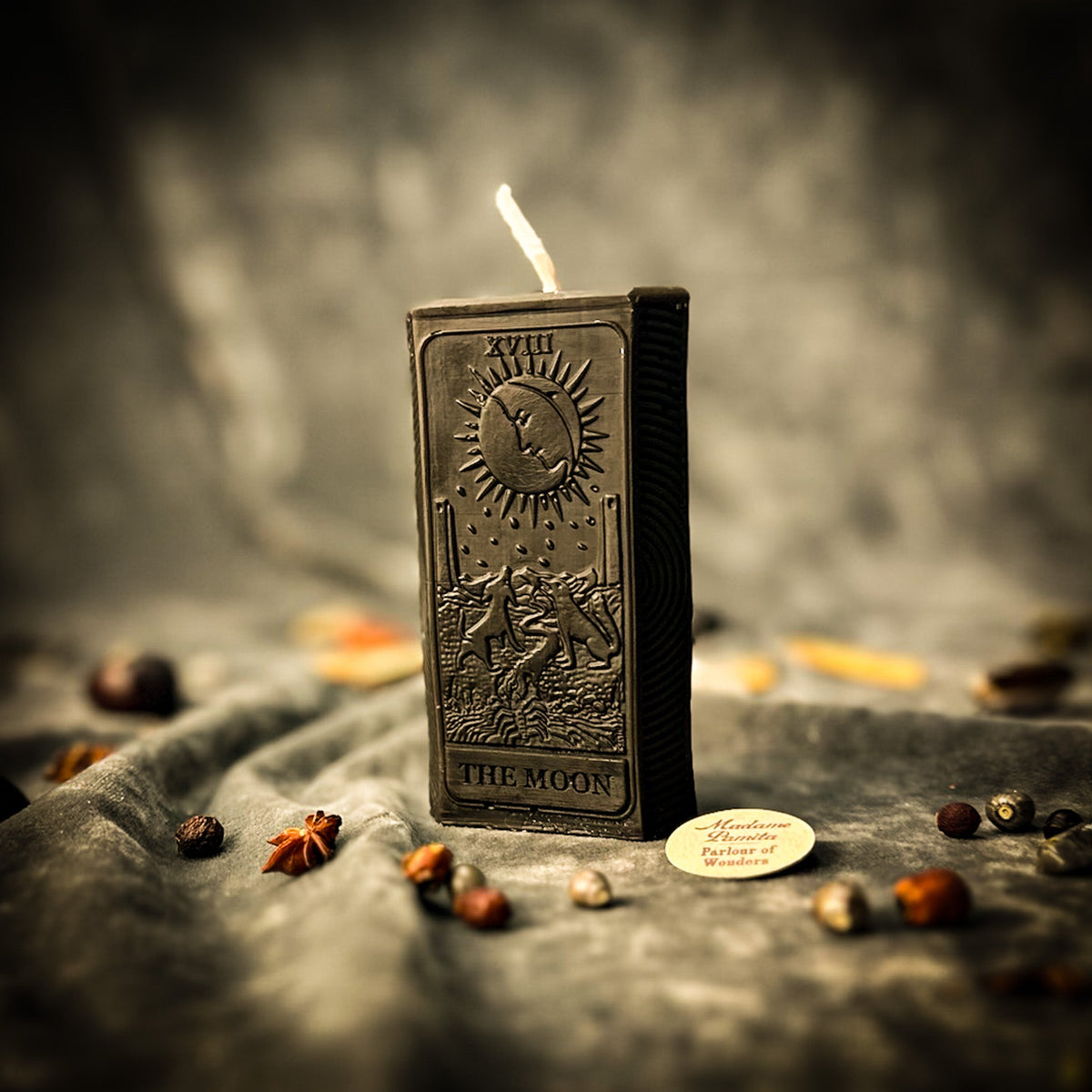 Black beeswax The Moon tarot card candle on gray cloth scattered with dried herbs and star anise
