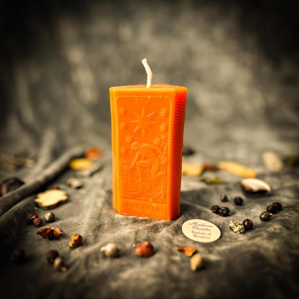 Orange beeswax The Star tarot card candle on gray cloth scattered with dried herbs and seeds