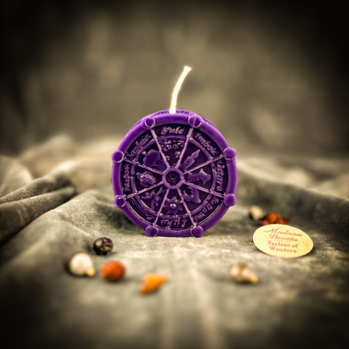 Purple Wheel of the Year candle on gray velvet, the eight sabbat names — Yule, Imbolc, Ostara, Beltane, Litha, Lammas, Mabon and Samhain — running around the rim.