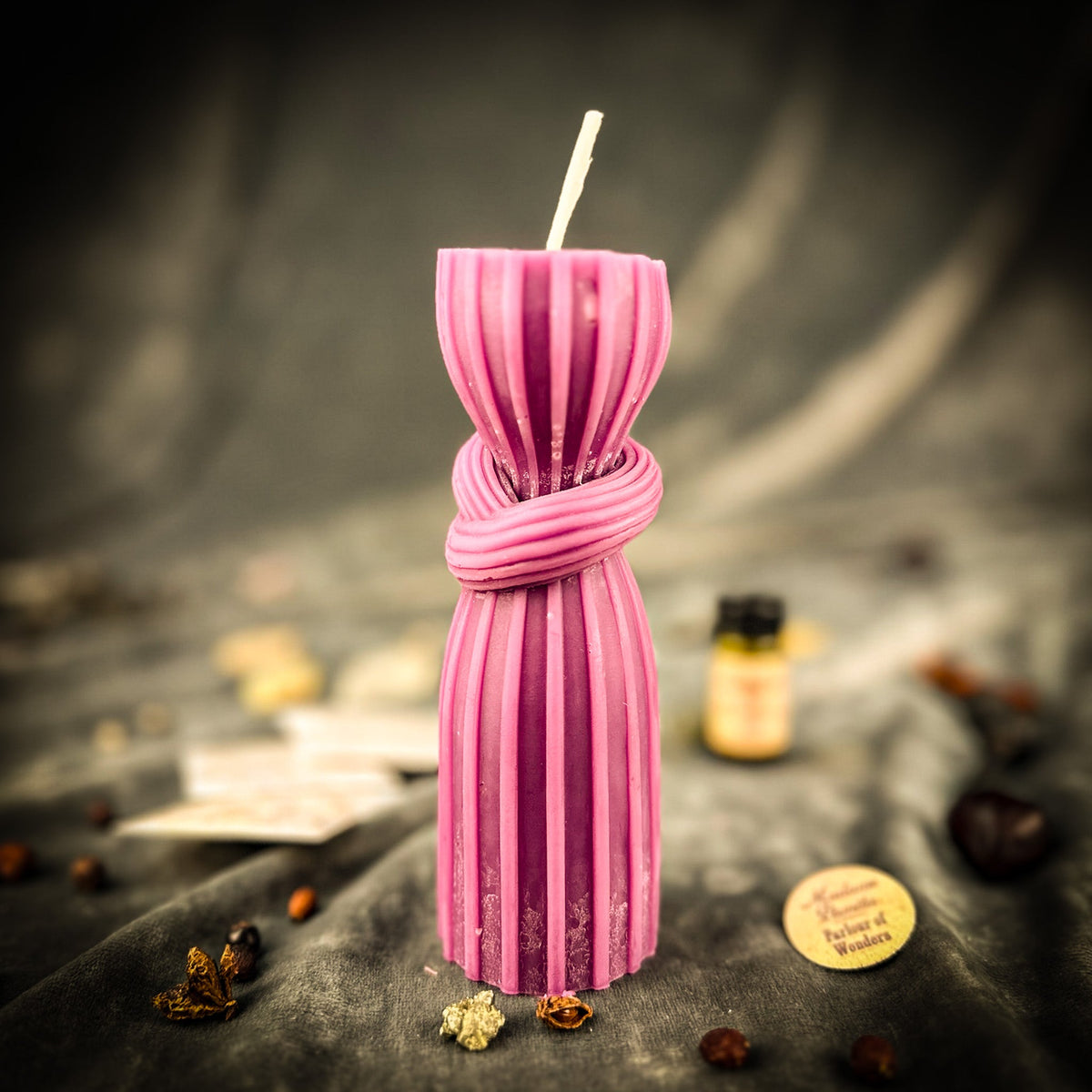 The pink beeswax knot candle standing on dark draped cloth, a ribbed column bound with a wax knot at its waist, oil vial and packets blurred behind