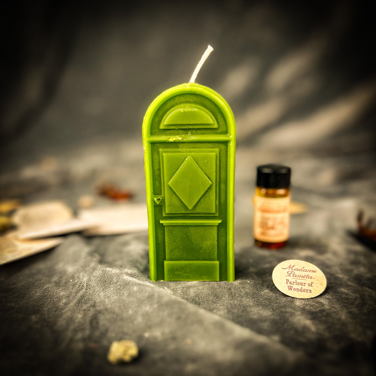Green door-shaped spell candle on gray velvet beside a small amber vial of dressing oil and a folded paper slip