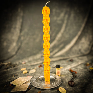 Golden yellow beeswax skull taper candle standing in a brass holder beside tarot cards and a vial of dressing oil