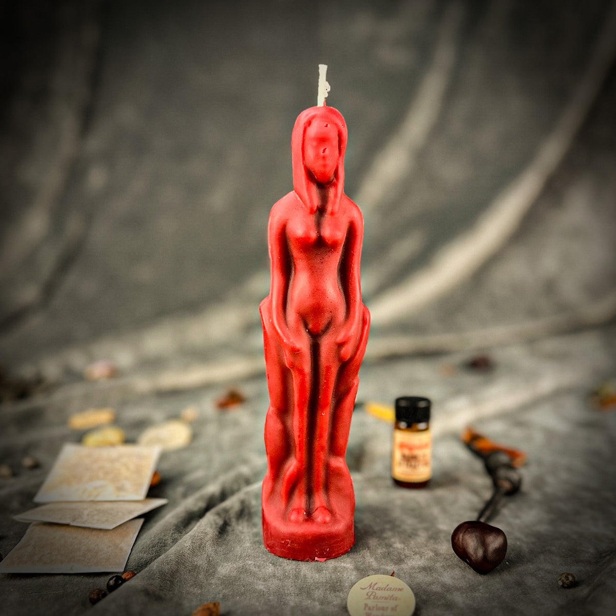 Red beeswax female figure candle standing on gray fabric with a vial of dressing oil, herb packets and the instruction sheet behind it