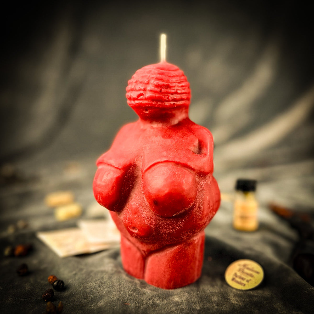 The red beeswax Willendorf goddess candle standing on dark draped cloth, its wick unlit, the oil vial and herb packets blurred behind