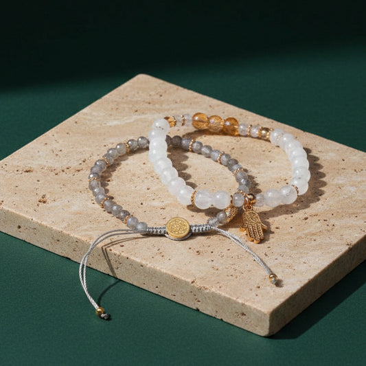 The two-bracelet wealth set on a travertine tile against dark green: citrine, moonstone and labradorite beads with a gold disc and a hamsa hand charm