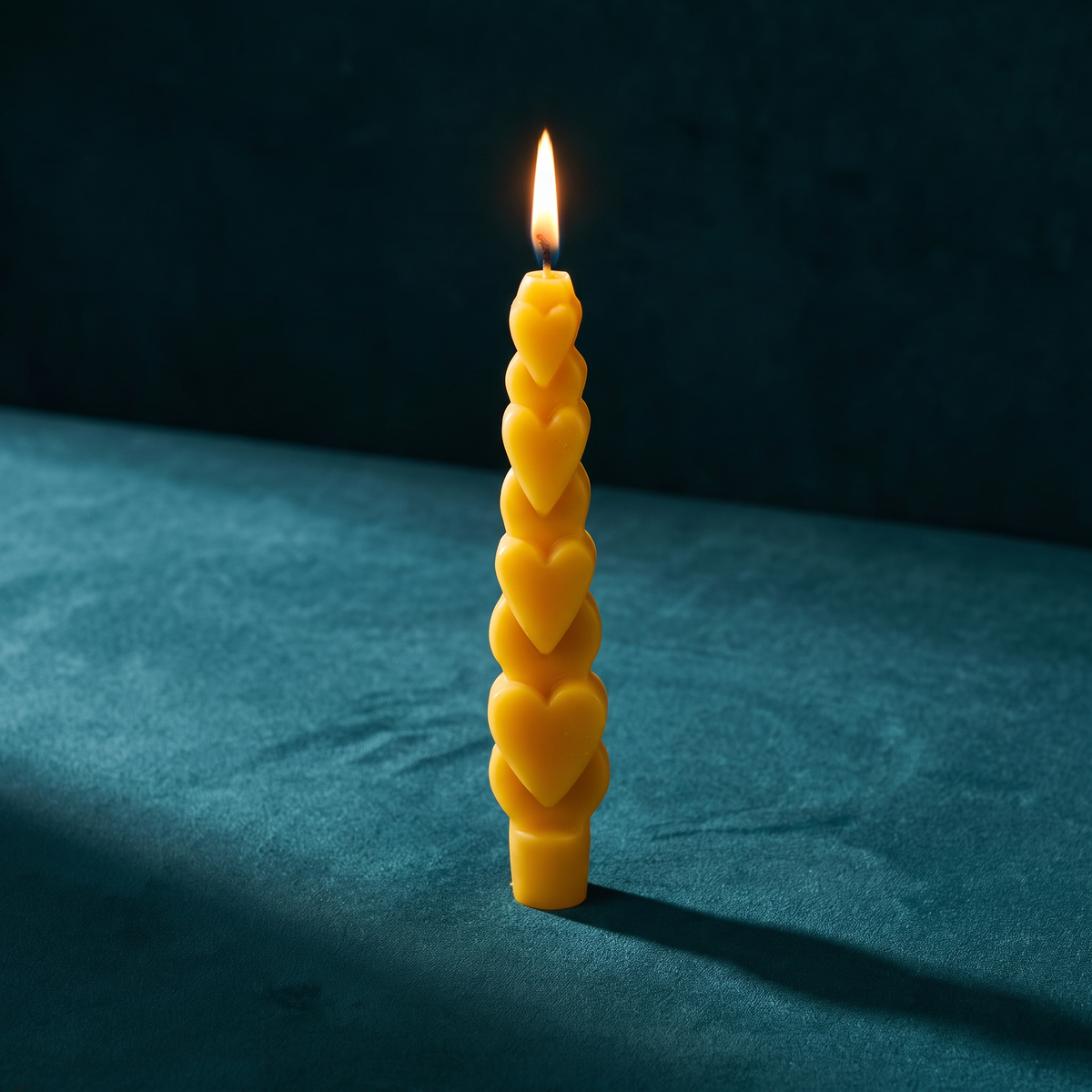 The same golden-yellow Love Spell Heart Taper alight, a steady flame at the wick, against teal velvet.