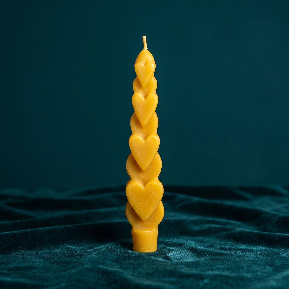 A single golden-yellow Love Spell Heart Taper standing unlit on teal velvet, the cast hearts spiraling up the column to a plain base socket.