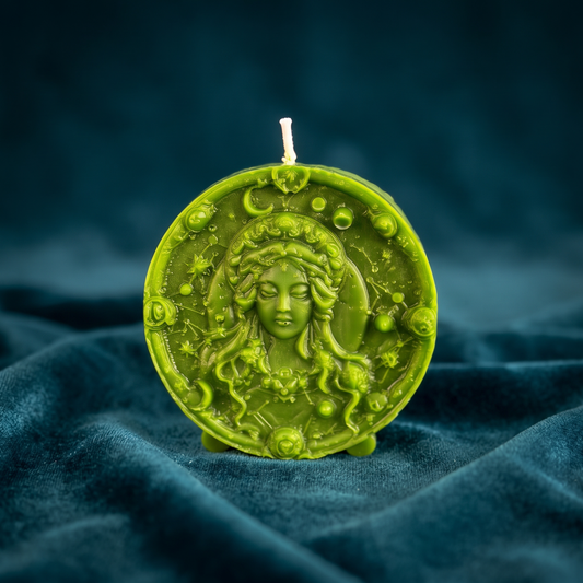 Olive-green Virgo candle on blue velvet: a round beeswax medallion standing on small feet, the Maiden's face in relief within a border of moons, stars and planets.