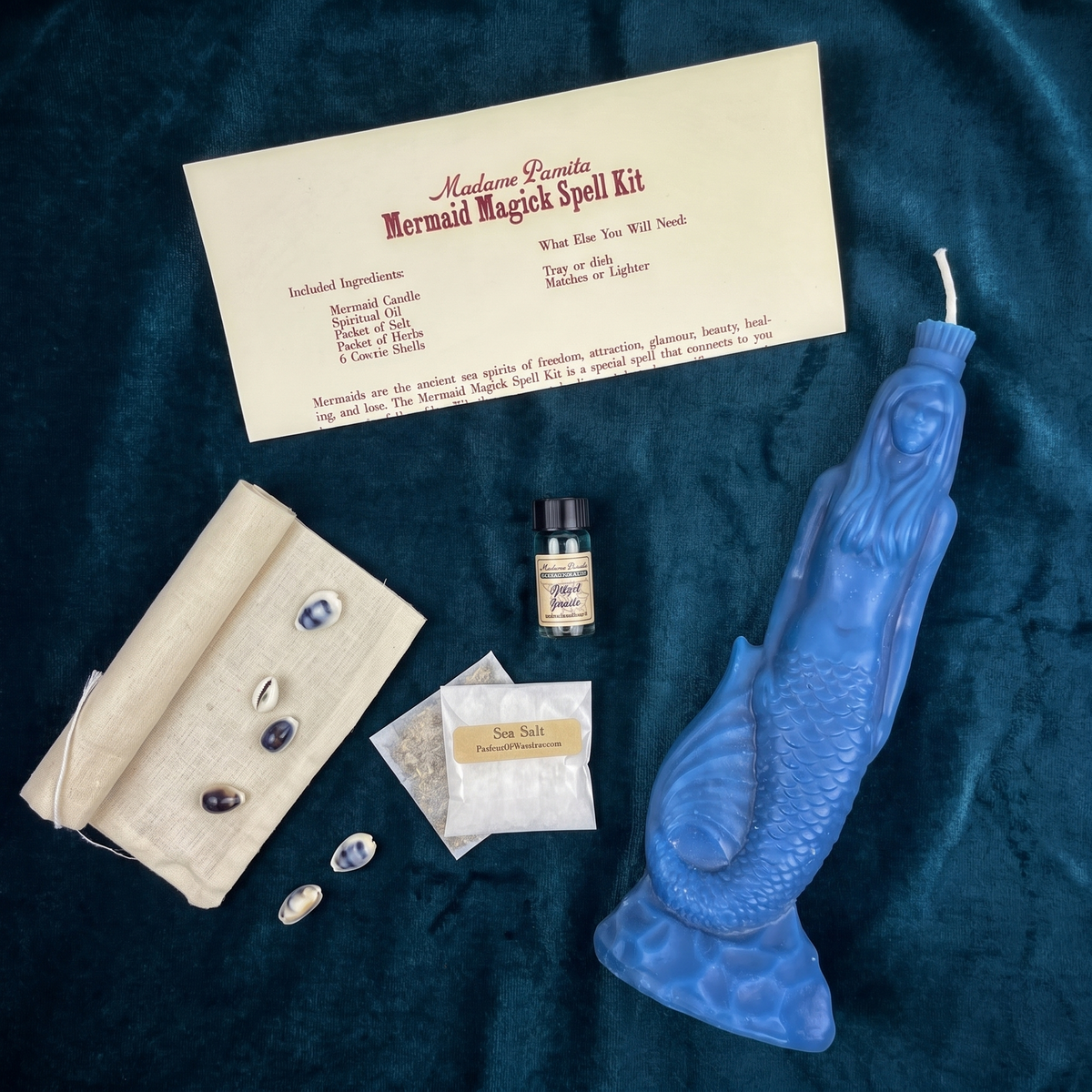 Mermaid Magick kit on teal velvet: a blue-lavender beeswax mermaid candle, the printed instruction card, a linen drawstring bag with cowrie shells, a bottle of spiritual oil and a sachet of sea salt.
