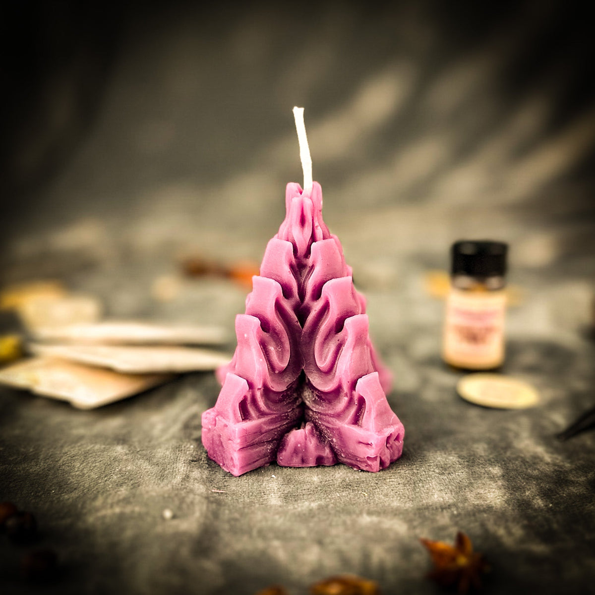 The mauve beeswax flame candle standing on dark draped cloth, its layered tongues of wax rising to an unlit wick, oil vial and herb packets blurred behind