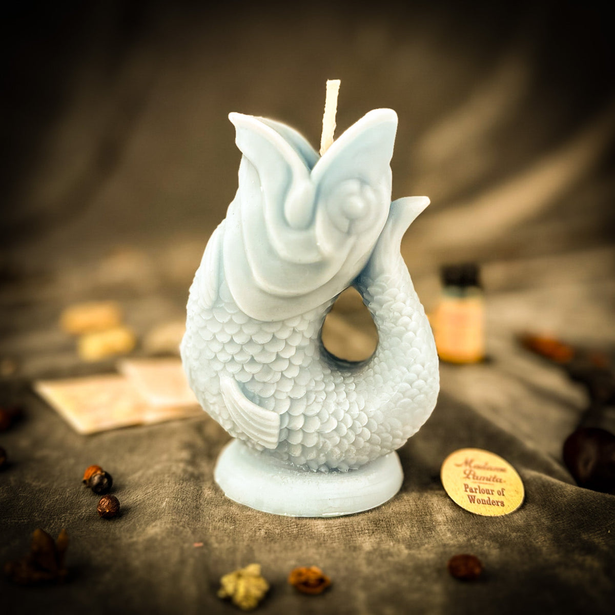 Pale blue fish-shaped candle on gray velvet with folded instruction papers, a small oil vial and scattered seeds