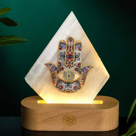 Selenite lamp with a diamond-shaped plate carrying a colorful Hamsa hand motif, lit from a rounded wooden base.