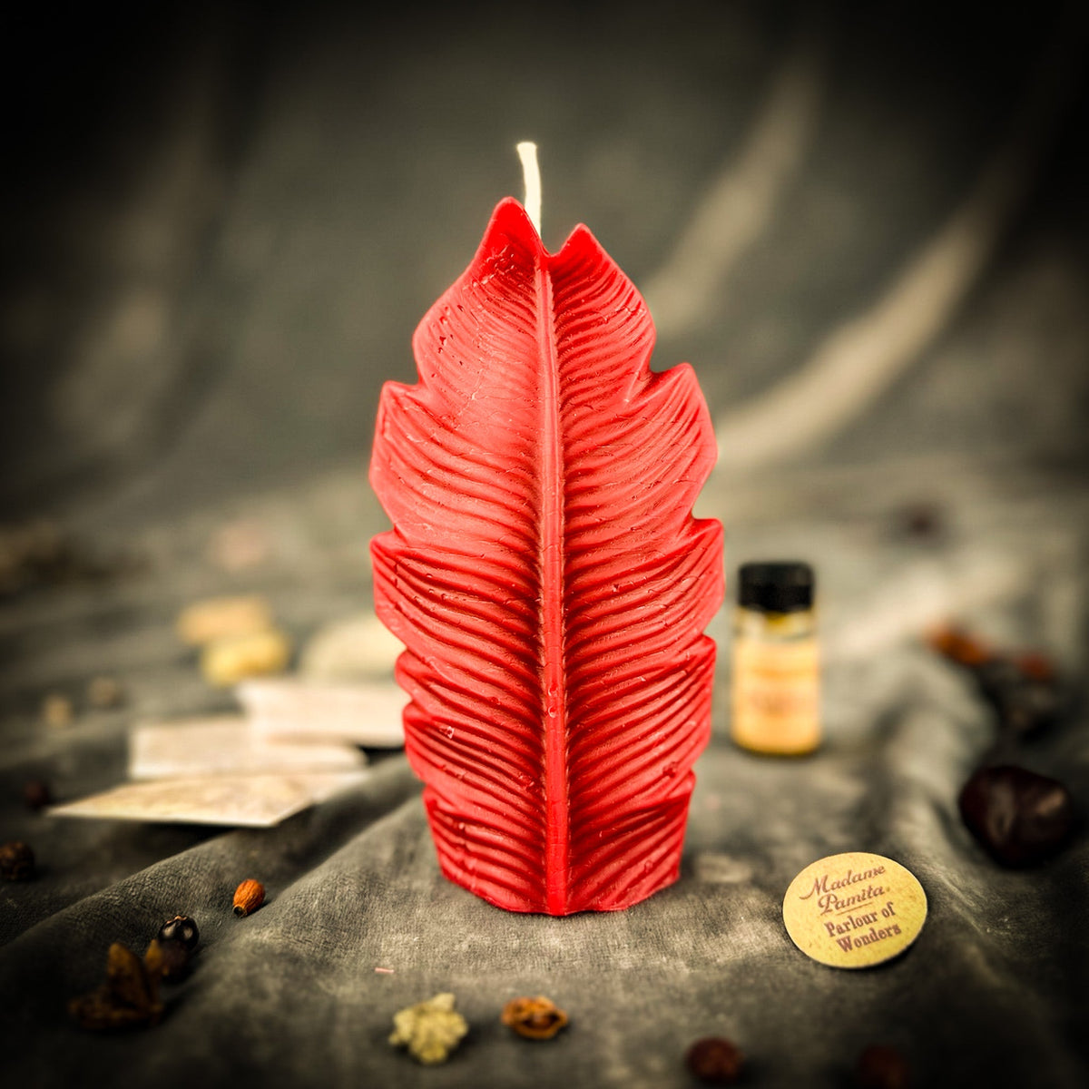 Red leaf-shaped candle on gray velvet with a small vial of dressing oil and folded instruction papers behind