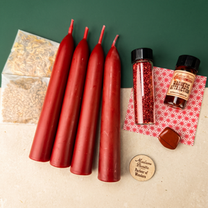 Attraction Candle Spell Booster Pack laid out on a green and cream backdrop: four red beeswax tapers, a vial of red glass glitter, a Magnetic Attraction ritual oil bottle, a red patterned card, two herb packets, a reddish-brown tumbled stone and a wooden Madame Pamita token from Parlour of Wonders.