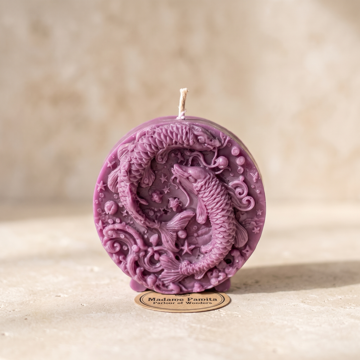 Mauve Pisces candle on a sand-colored surface: a round beeswax medallion on small feet, two fish circling head to tail among stars, bubbles and waves, the Madame Pamita label at its foot.