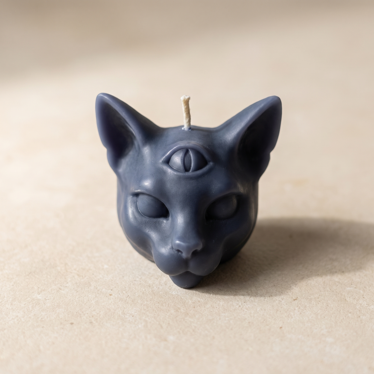 Slate-blue beeswax cat head candle on a sand-colored surface, a vertical third eye molded on the forehead and the wick rising between the ears.