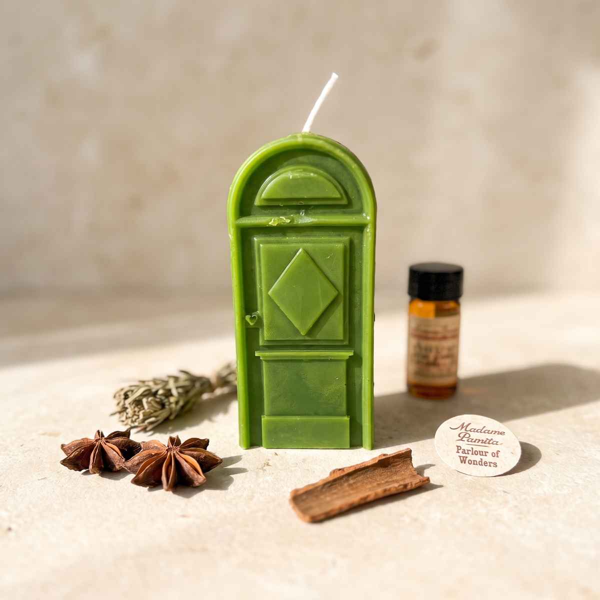 Green door-shaped spell candle standing on pale stone with a small vial of dressing oil, a sage bundle, star anise and a cinnamon stick