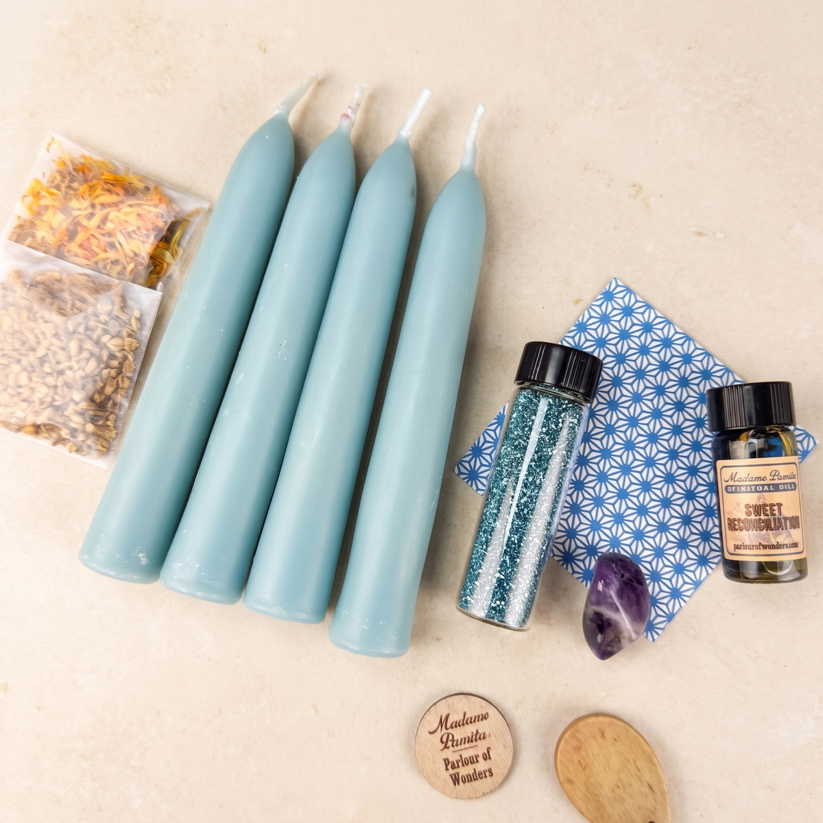 Reconciliation Candle Spell Booster Pack on pale stone: four dusty blue beeswax tapers, a vial of teal glass glitter, a Sweet Reconciliation ritual oil bottle, a blue star-patterned card, two herb packets, a purple tumbled stone and a wooden Madame Pamita token from Parlour of Wonders.