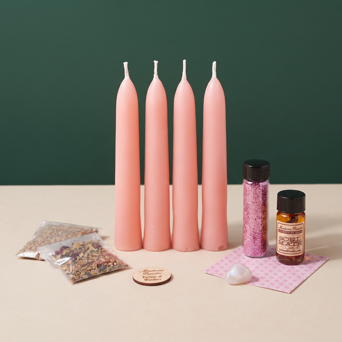 Romance Candle Spell Booster Pack on a green and cream backdrop: four pink beeswax tapers, a vial of pink glass glitter, a Soul Mate ritual oil bottle, a pink patterned card, two herb packets, a pale opalescent tumbled stone and a wooden Madame Pamita token from Parlour of Wonders.