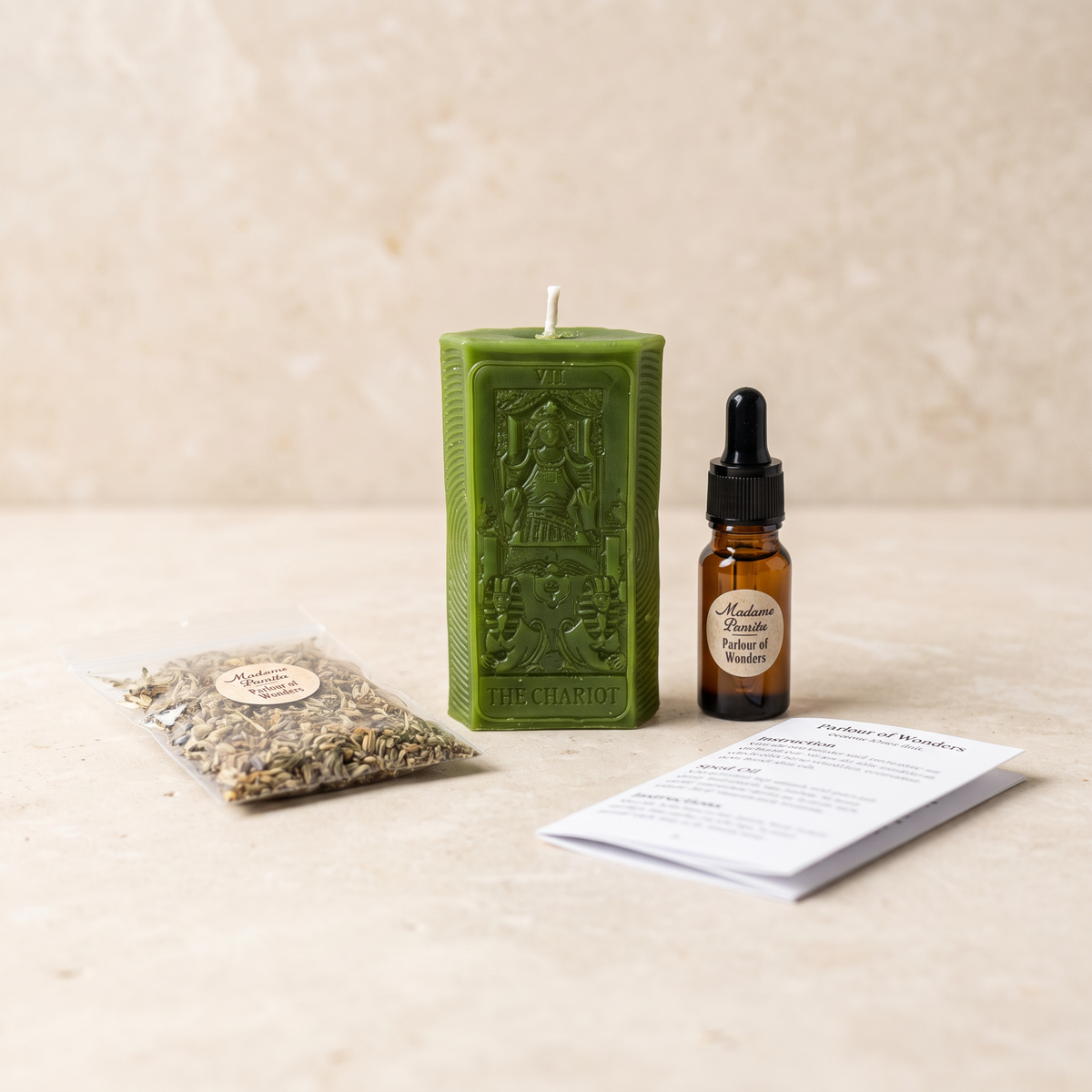 The Chariot candle spell kit laid out on stone: a green beeswax tarot-card candle with a dropper bottle of dressing oil, a pouch of dried herbs and the printed instruction sheet