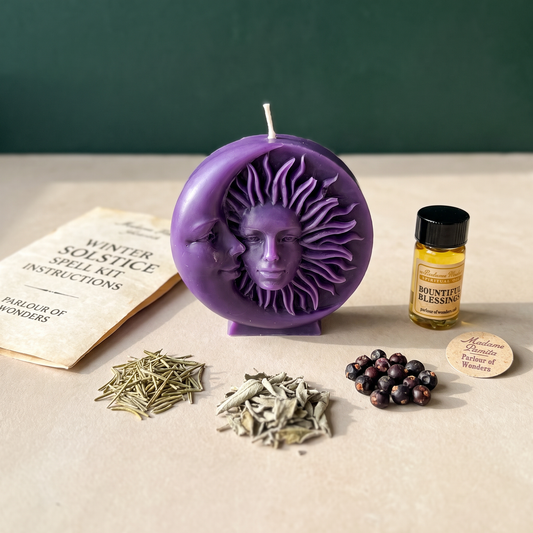 Purple sun-and-moon disc candle beside the printed Winter Solstice spell kit instructions, two heaps of dried herbs, juniper berries and a vial of Bountiful Blessings oil