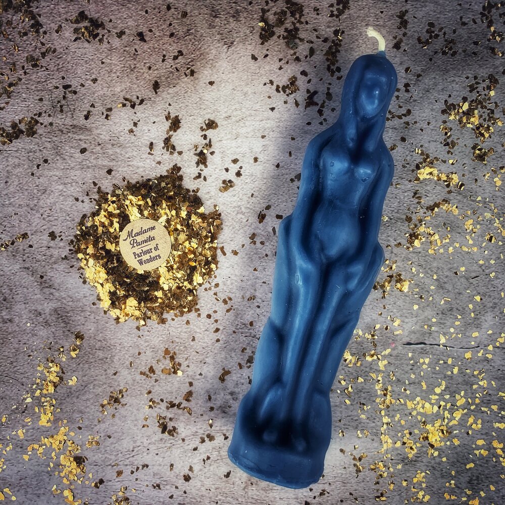 Blue beeswax female figure candle on gray cloth scattered with crushed herbs and gold leaf