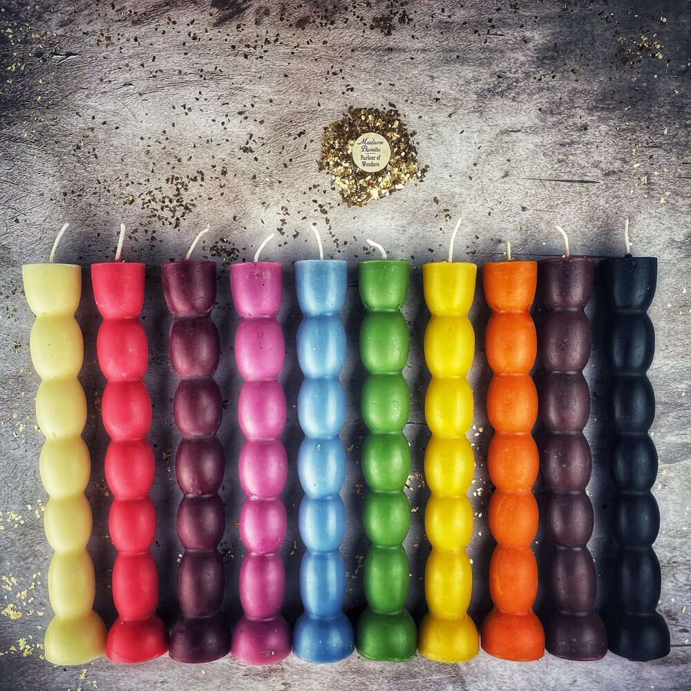 All ten seven knob candles laid flat in a row on gray stone — cream, red, plum, magenta, blue, green, yellow, orange, aubergine and black — wicks at the top.