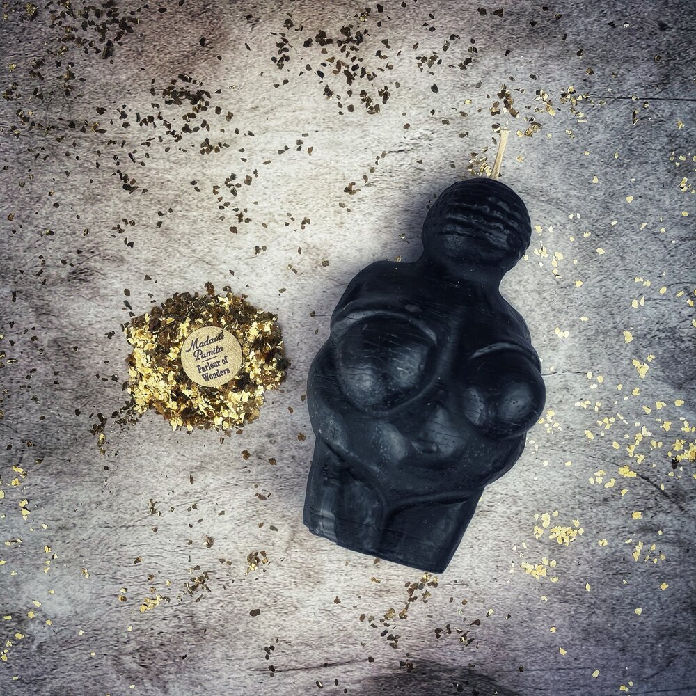 The black beeswax goddess candle lying on weathered gray stone among scattered dried herbs and gold flakes, the round maker's seal beside it