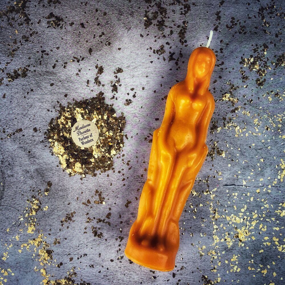 Orange beeswax female figure candle on gray cloth strewn with herbs and gold leaf