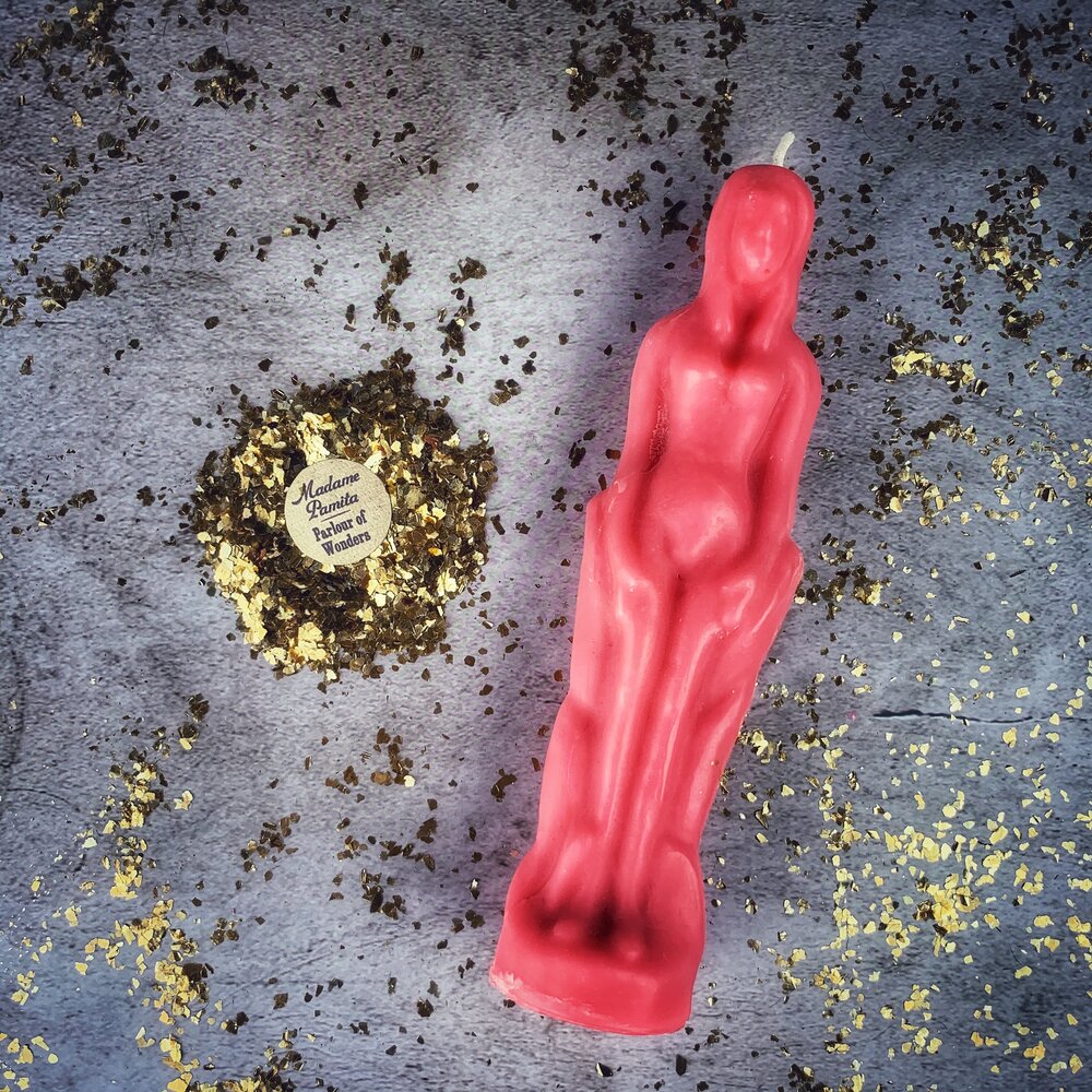 Pink beeswax female figure candle lying on gray cloth strewn with dried herbs and gold flecks