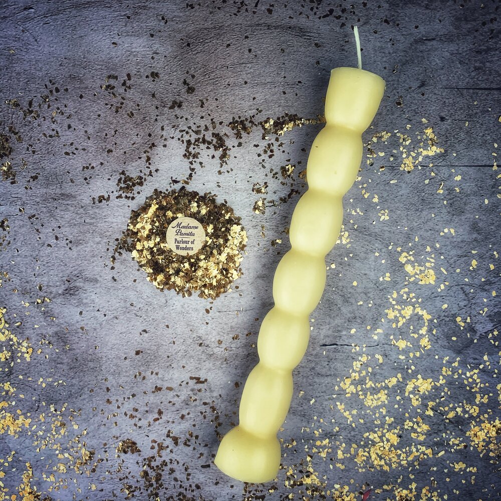 Cream-white seven knob candle lying on gray stone beside the round Parlour of Wonders label and scattered gold leaf.